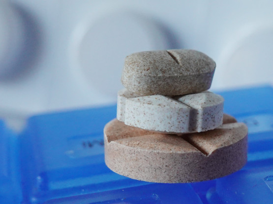 Drei unterschiedlich große, braun bis beige gefärbteTabletten, die aufeinander gestapelt auf einem blauen Tablettenkästchen liegen. Im Hintergrund eine Blisterverpackung.